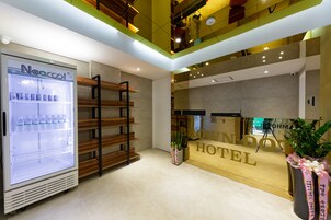 Un área de recepción de hotel con un refrigerador marca Neocool, estanterías de madera y un mostrador de recepción con un cartel que dice 'BLOW N'DOX HOTEL'.