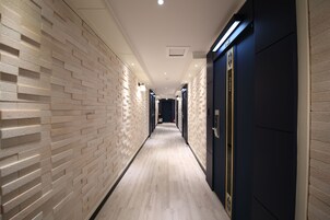 Un pasillo largo y estrecho, con una pared de ladrillo claro en un lado y una pared azul oscuro en el otro. El suelo es de madera clara y el pasillo está bien iluminado con luces de techo.