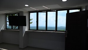 A room with a bed, a TV mounted on the wall, and large windows with a view of the landscape.