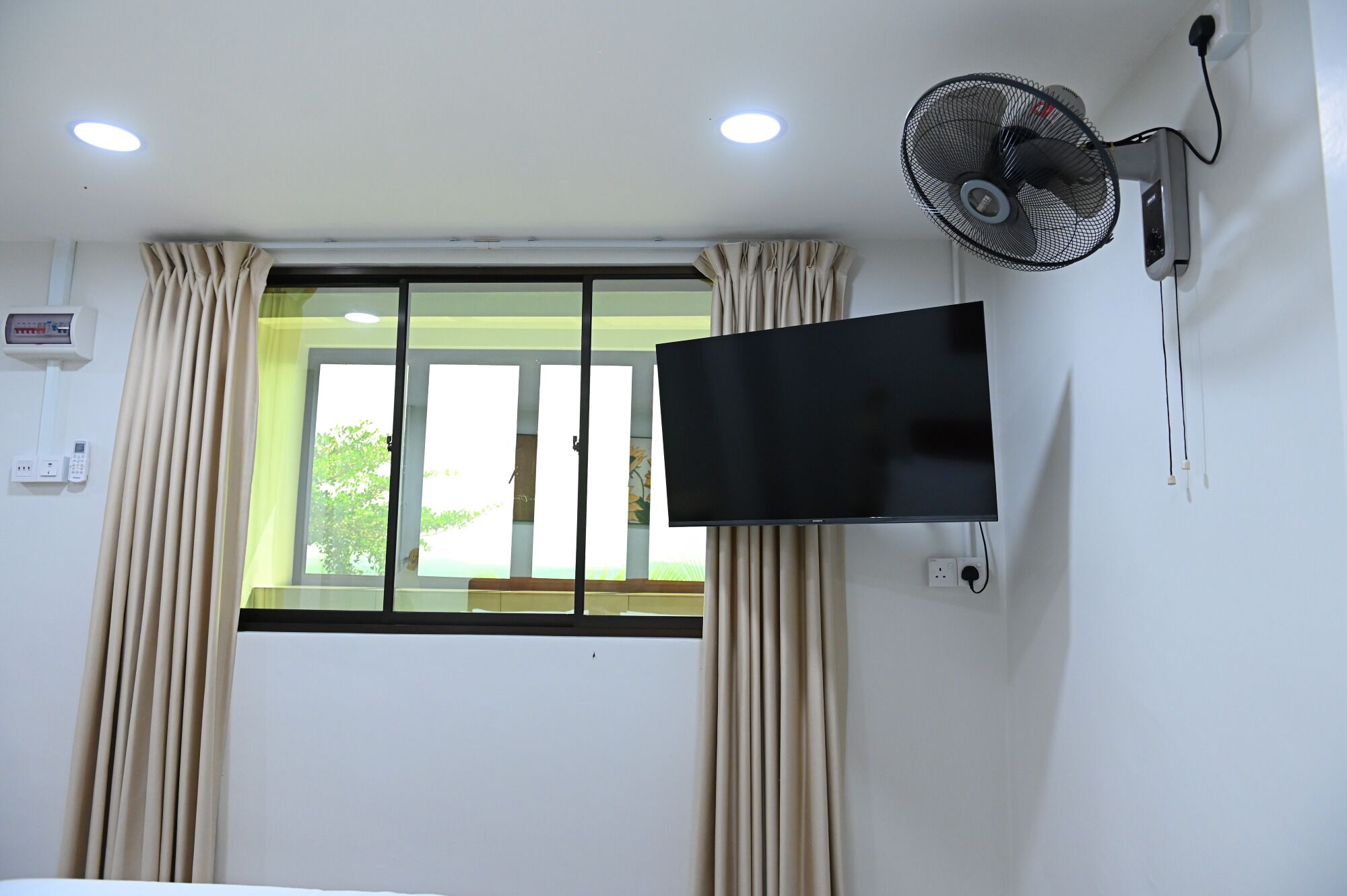 A room with a window, curtains, a mounted TV, a wall-mounted fan, and a ceiling light.