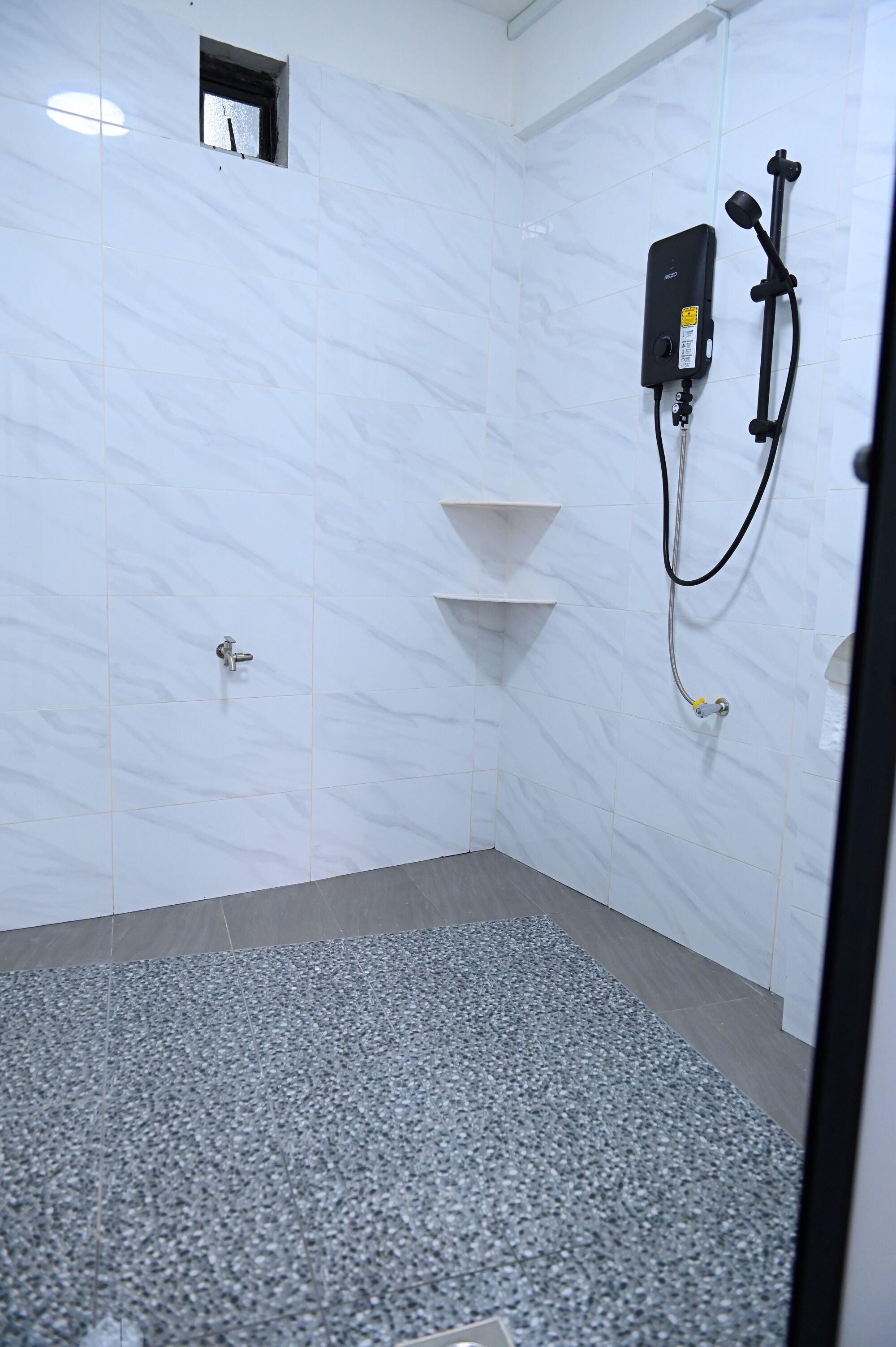 A bathroom with white tiled walls, a shower with a handheld showerhead, and a small window.