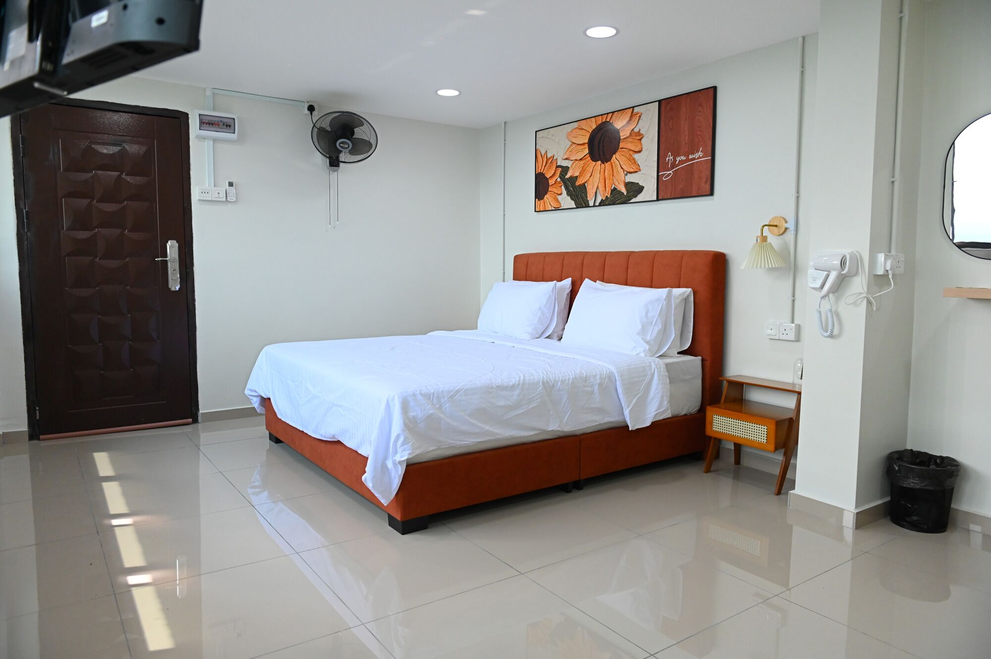 Habitación de hotel con cama, ventilador de pared, cuadro, mesita de noche, secador de pelo y cesto de basura.