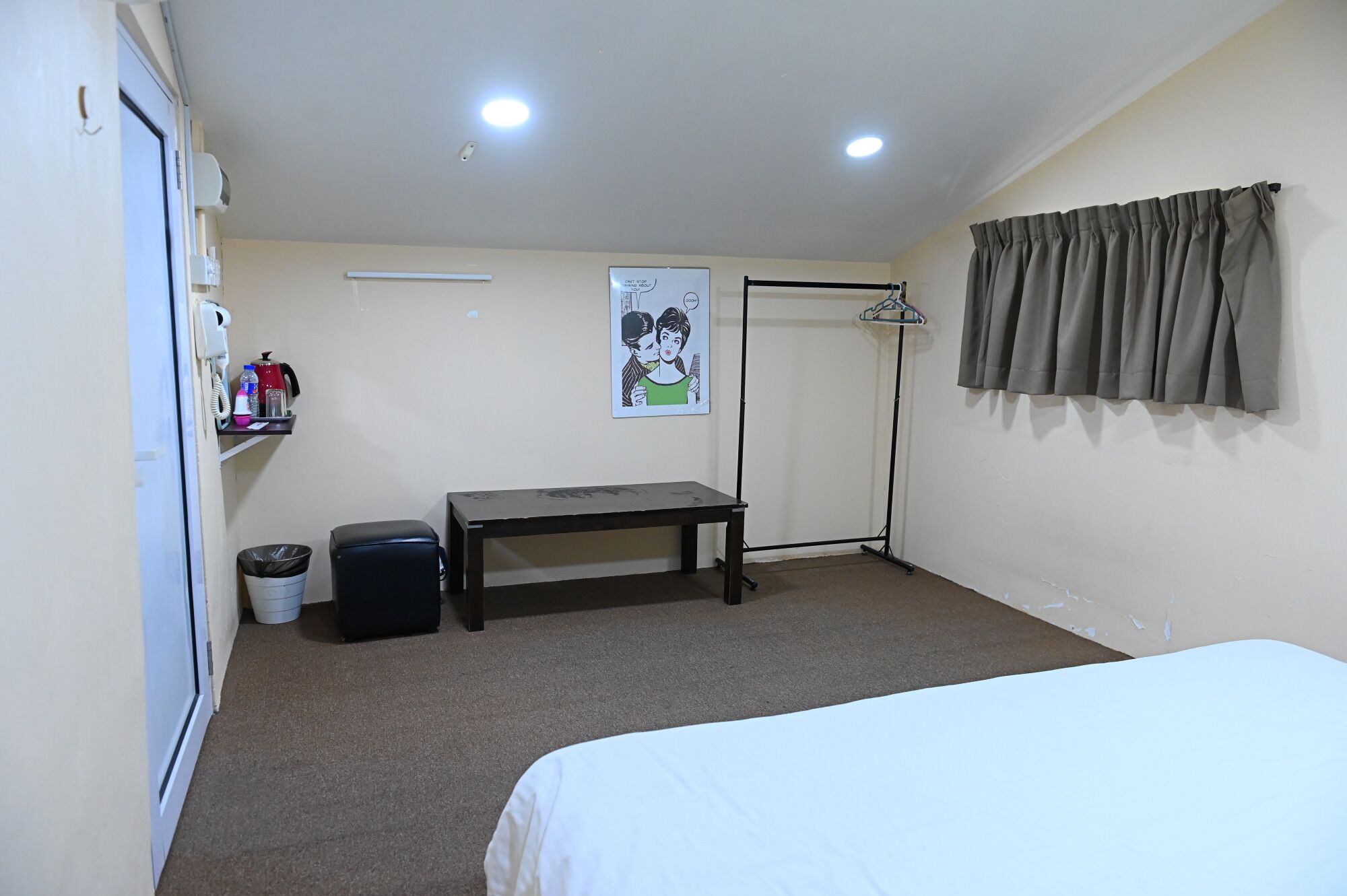 Una habitación de hotel pequeña con cama, escritorio, silla, cubo de basura, perchero y un cartel en la pared.