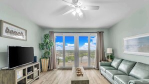 Una sala de estar con televisor, sofá, mesa de centro y vista al exterior.