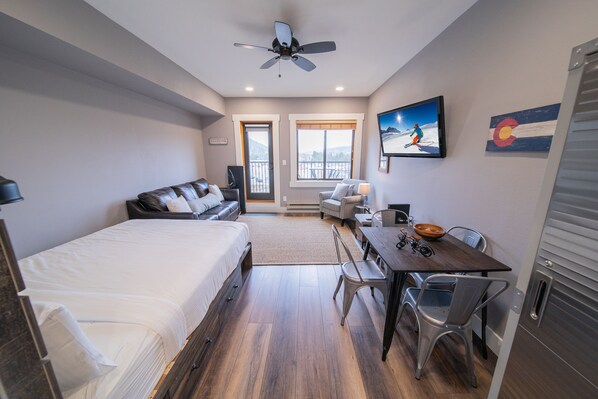 A modern hotel room with a large bed, a desk with a chair, a ceiling fan, and a view of the outdoors through the windows. - Updated Studio | Balcony & Hot Tub, Firepit (Keystone)