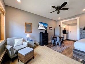 A modern hotel room with a sofa, ottoman, ceiling fan, TV, dining table, chairs, and a kitchen area. - Updated Studio | Balcony & Hot Tub, Firepit (Keystone)