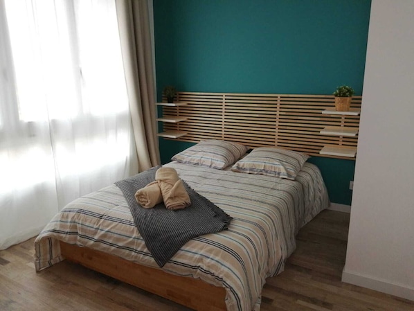 A neatly made bed with striped bedding and a rolled towel, wooden headboard, and a shelf with a plant.