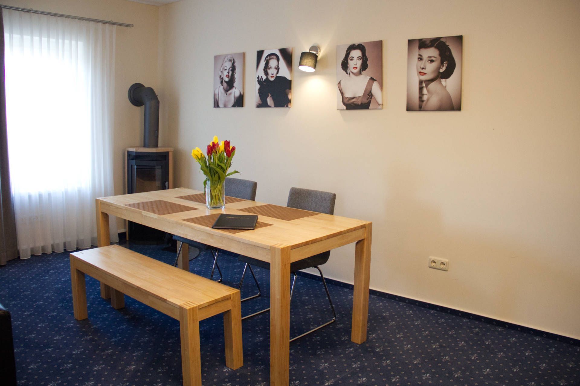 A hotel room with a wooden dining table, a vase of flowers, and four framed pictures on the wall.
