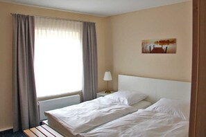 A hotel room with two beds, a window with curtains, a nightstand with a lamp, and a painting on the wall. - Am Wiesenblick - Müritz apartment (Röbel/Müritz)