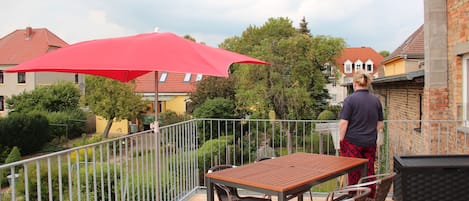 A balcony with a wooden table and chairs, a red umbrella, and a brick building in the background.