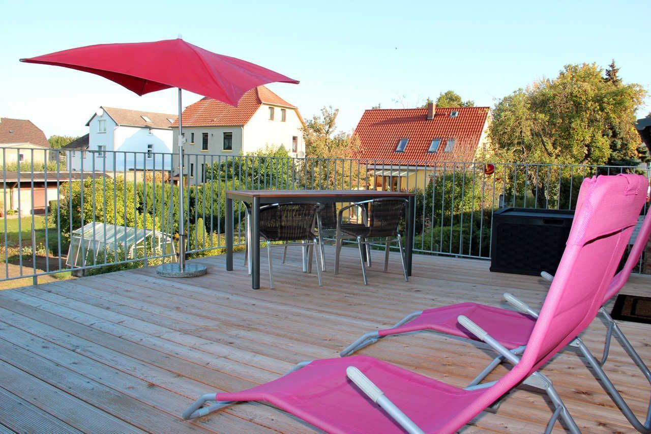 A wooden deck with a table and chairs, a red umbrella, and a pink chair.