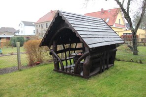A rustic wooden structure with a sloped roof, a bench, and a table, surrounded by a grassy area and a fence. - Holiday ivy - Müritz apartment (Röbel/Müritz)