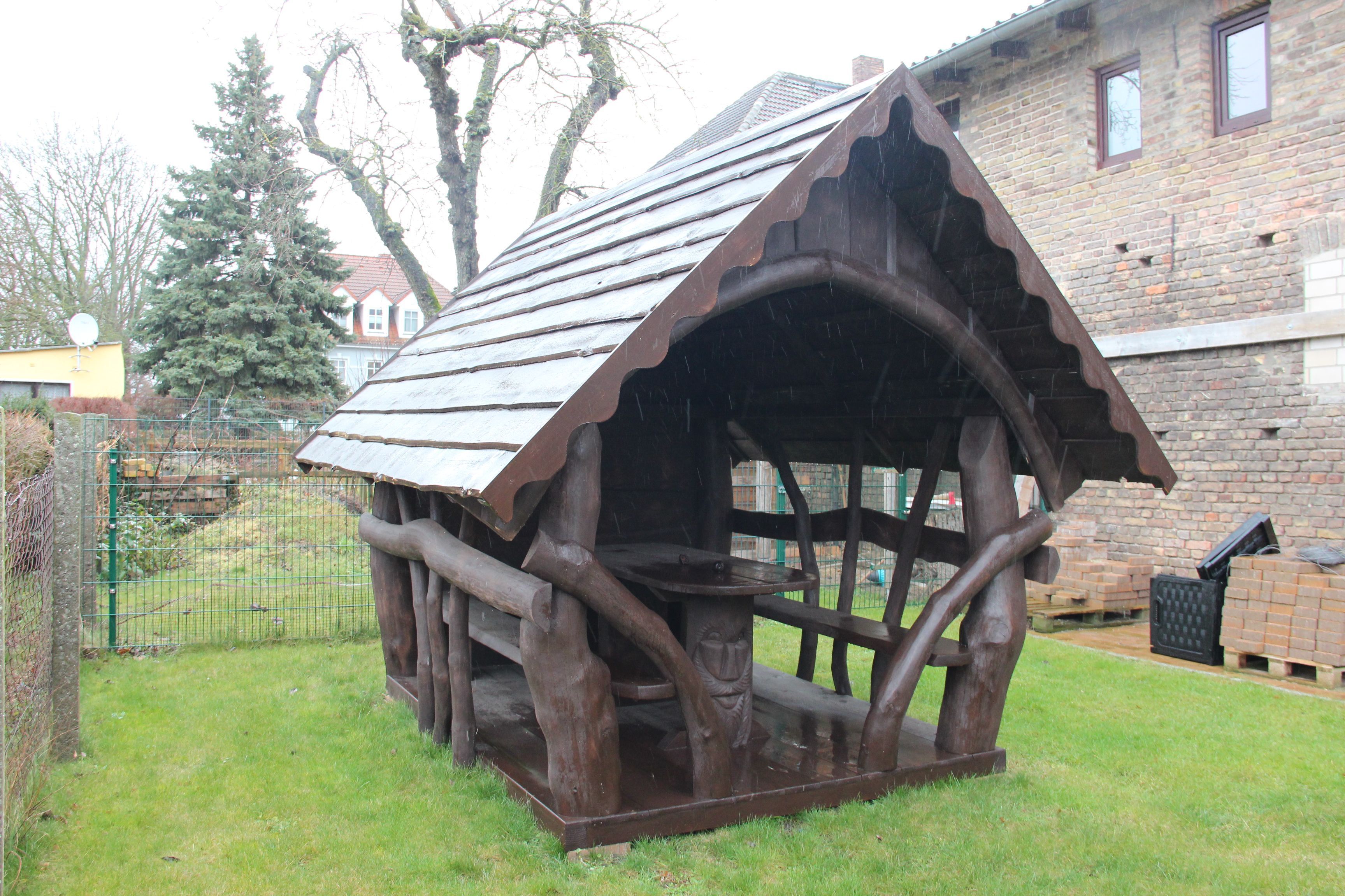 A rustic wooden structure with a sloped roof, surrounded by a grassy area and a brick building in the background.