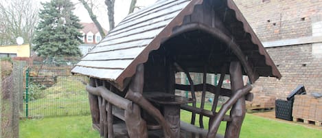 A rustic wooden structure with a sloped roof, surrounded by a grassy area and a brick building in the background.