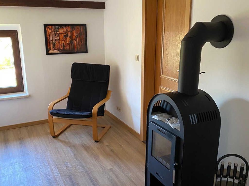 A room with a wooden floor, a black upholstered chair, a black wood-burning stove, a wooden door, and a framed picture on the wall.