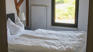 Una cama con ropa de cama blanca, una lámpara de noche y una ventana con vistas a la vegetación.