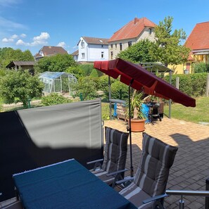En terrasse med et bord og stole, en rød parasol og et drivhus i baggrunden.