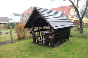 A rustic wooden structure with a sloped roof, situated in a grassy area with a fence and houses in the background.