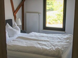 A bed with white bedding, a bedside lamp, and a window with a view of greenery.