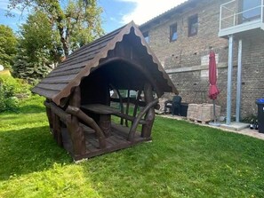 A rustic wooden structure with a sloped roof, possibly a gazebo or playhouse, situated in a grassy area with a brick building in the background. - Tenne 1 - Müritz apartment (Röbel/Müritz)