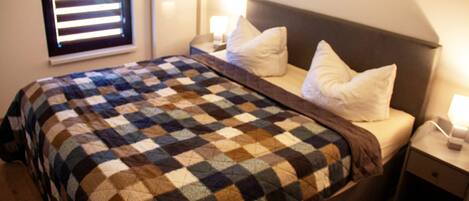 A neatly made bed with a checkered blanket, a bedside table with a lamp, and a window with blinds.