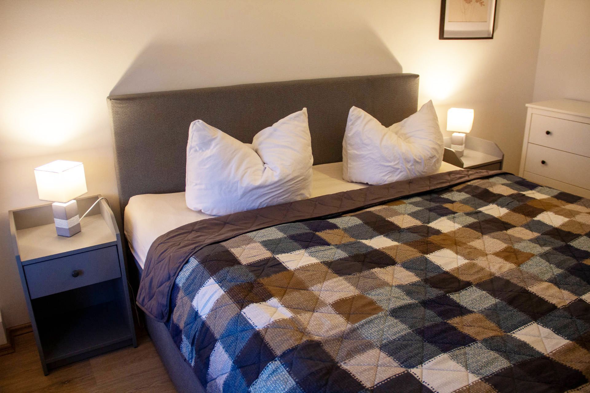 A neatly made bed with a checkered blanket, two white pillows, and a bedside lamp on a nightstand.