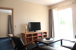 Une chambre d’hôtel équipée d’une télévision posée sur un meuble en bois, d’un canapé, d’une table basse et d’une porte-fenêtre coulissante donnant sur un balcon.