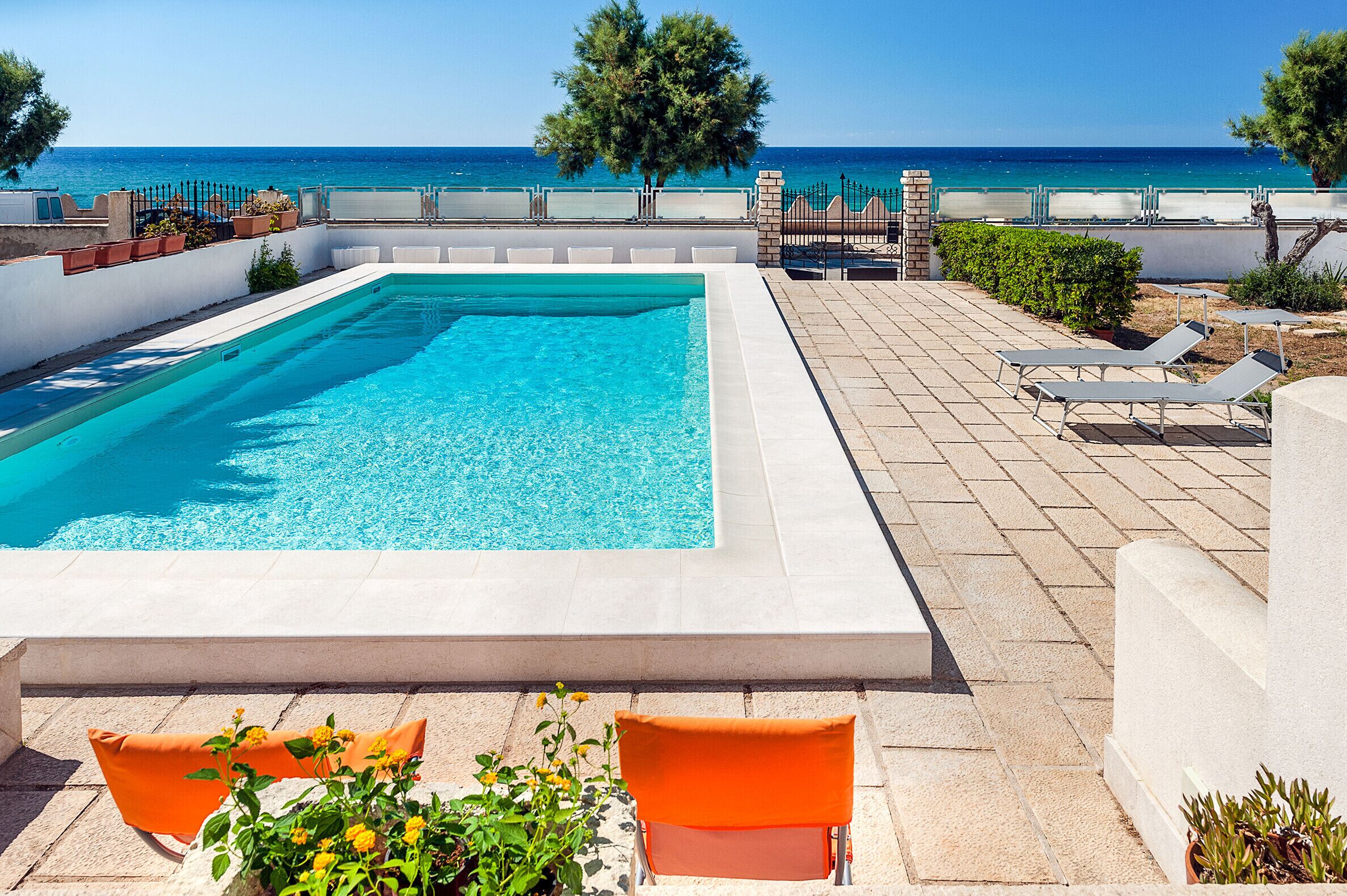 A rectangular swimming pool with a clear blue water, surrounded by a tiled deck with lounge chairs and potted plants.