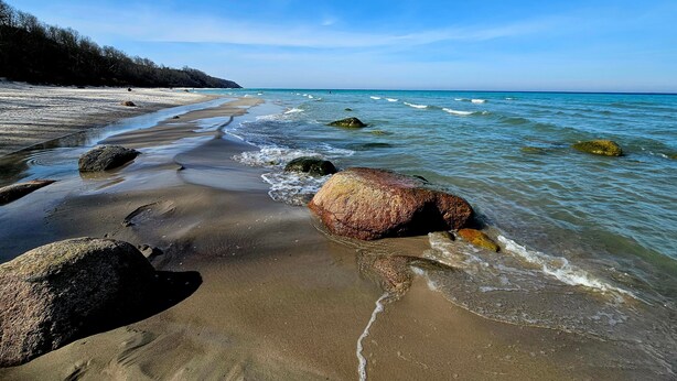 Een strand met helderblauw water, grote rotsen en een zandstrand.