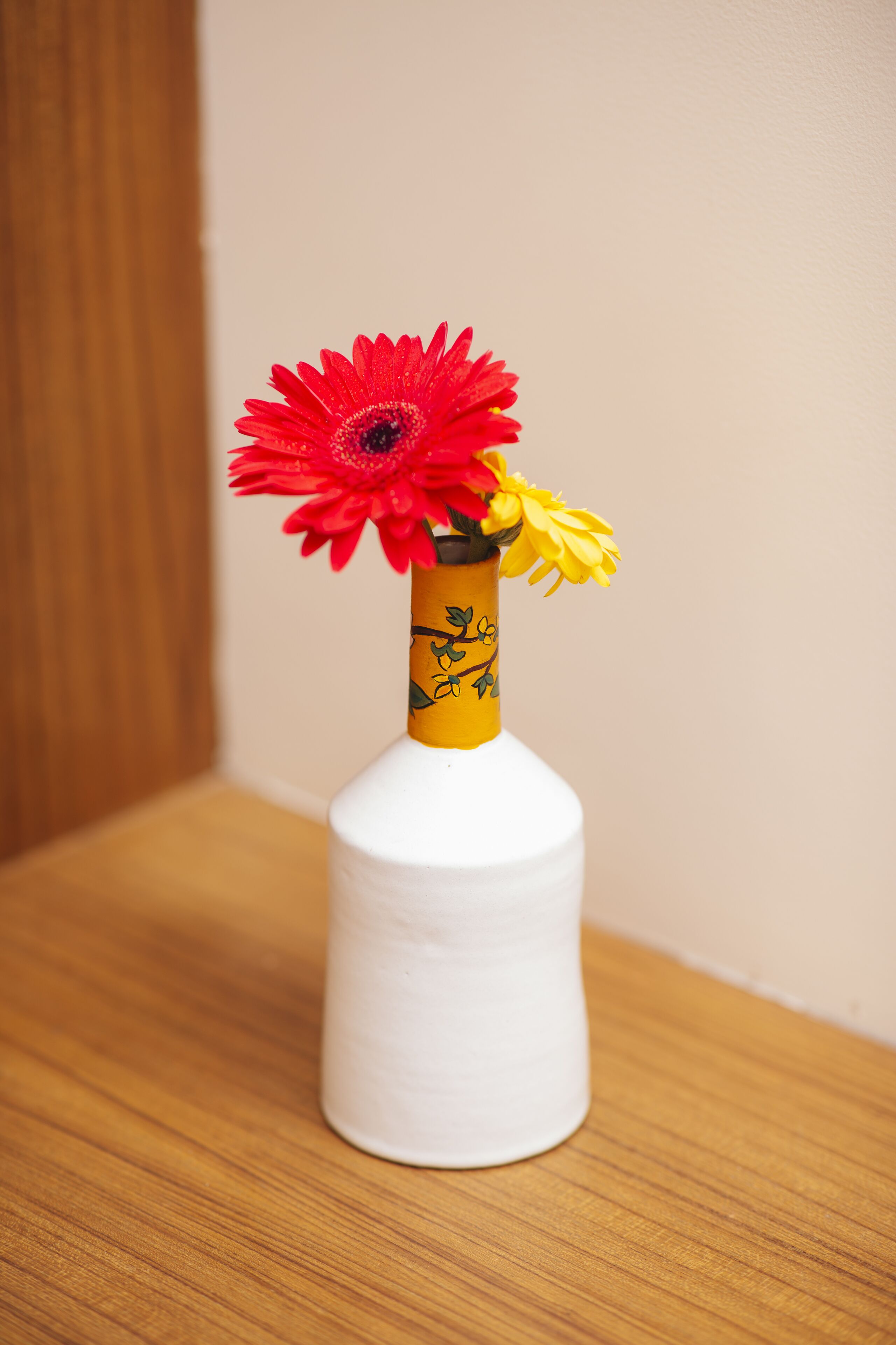 A white vase with a yellow band and floral design, containing red and yellow flowers, placed on a wooden surface.