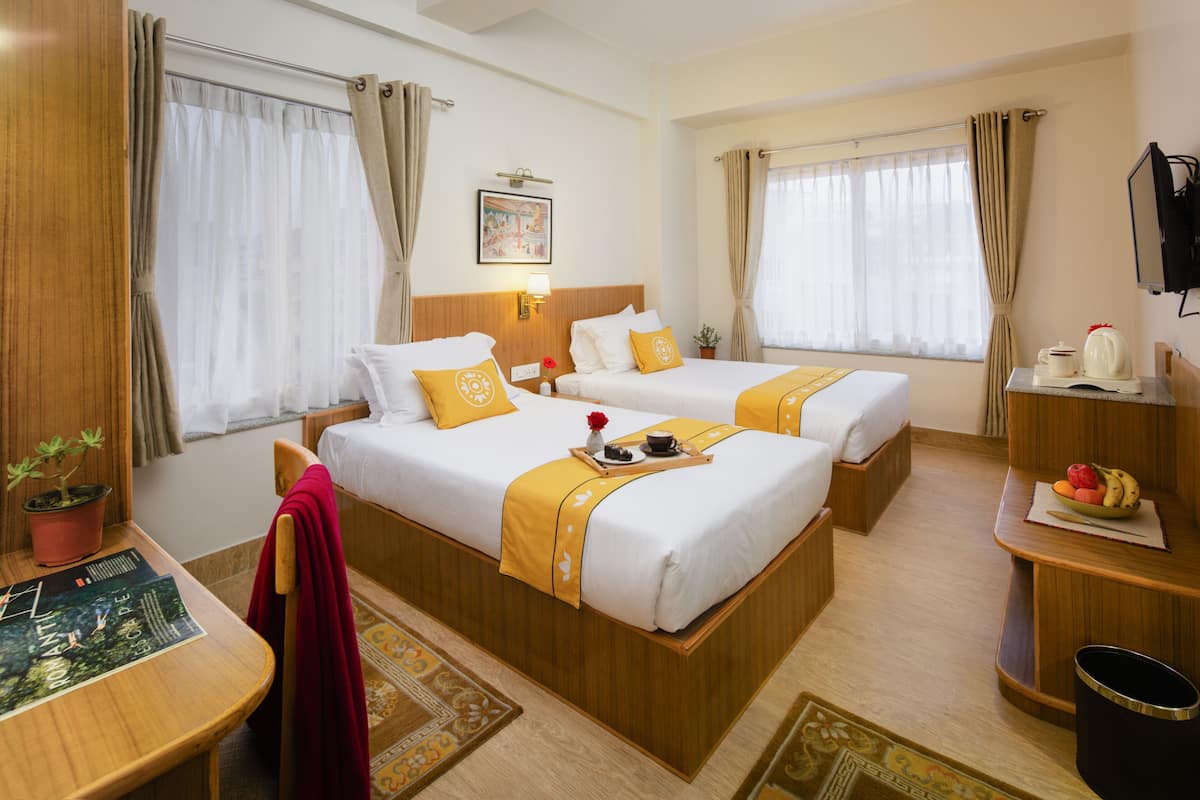 A hotel room with two beds, a desk, a TV, and a small table with a fruit bowl.