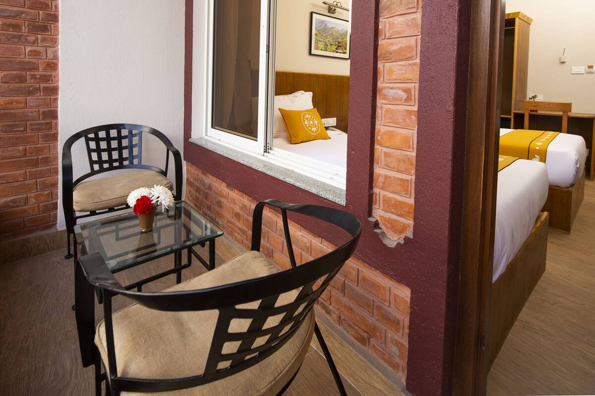 A hotel room with a brick wall, a window with a bed, a small table with chairs, and a wooden desk.