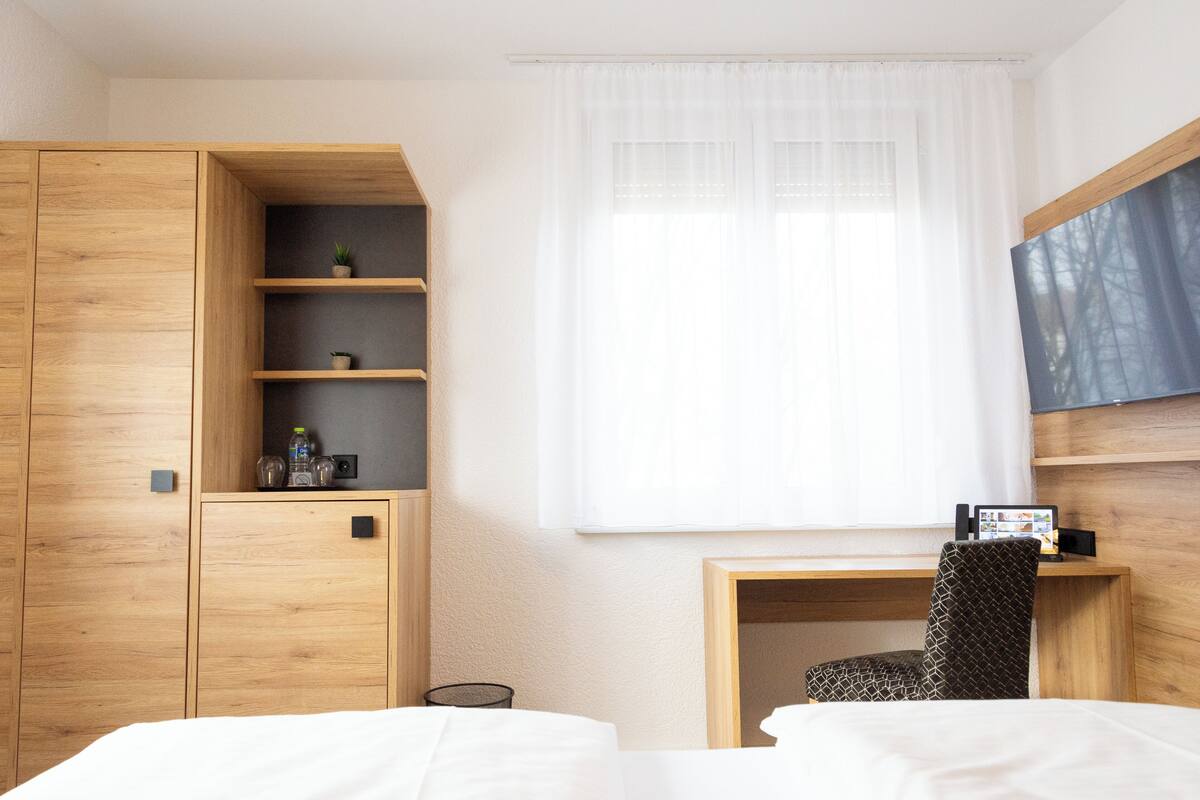 Una habitación de hotel con un armario de madera, un escritorio con silla, una cama y un ventanal con cortinas translúcidas.