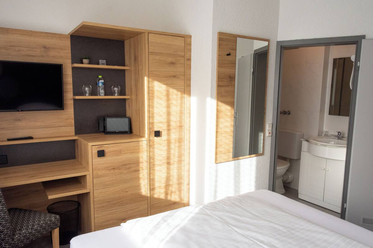 Una habitación de hotel con cama, televisor, escritorio con computadora, espejo y baño con lavamanos e inodoro.