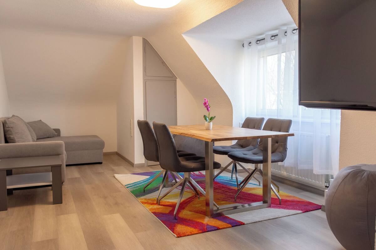 Una sala de estar moderna con comedor, que incluye un sofá, una mesa con sillas, una alfombra colorida y un televisor montado en la pared.
