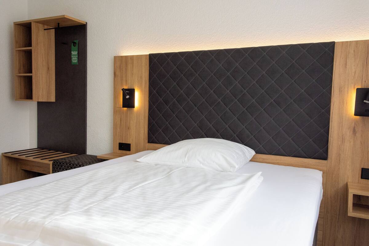Una cama bien hecha con un cabecero acolchado, una mesita de noche de madera y un pequeño estante con un letrero verde.