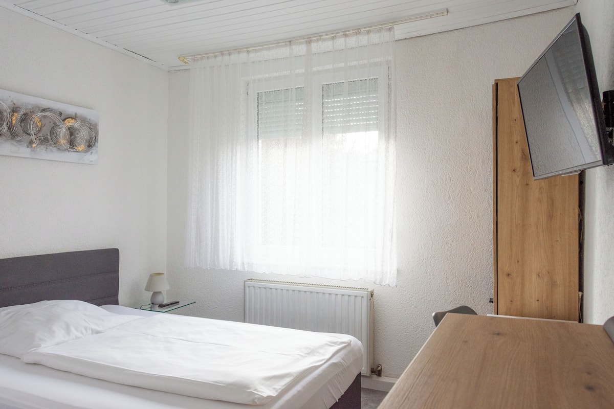Un dormitorio con cama, una mesita de noche con lámpara, una ventana con persianas, un radiador y un televisor montado en un panel de madera.
