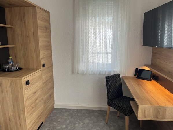 Une pièce de taille modeste, avec un bureau en bois, une chaise, un téléviseur fixé au mur et une fenêtre ornée de rideaux légers.