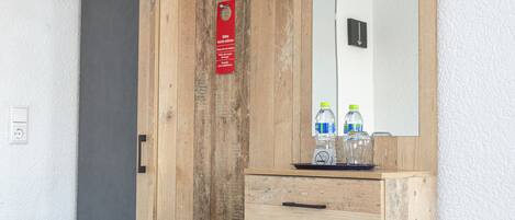 A room with a wooden wardrobe, a small safe, a mirror, and a shelf with bottled water.