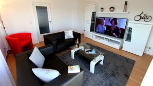 Un salon moderne avec un canapé noir, un fauteuil rouge et un téléviseur diffusant une émission.