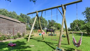 A wooden swing set, a hammock, a red toy car, a blue tricycle, and a horse in a grassy yard with a wooden fence and trees in the background. - Apartment West - Ferienhof Deich in sight (Esens)