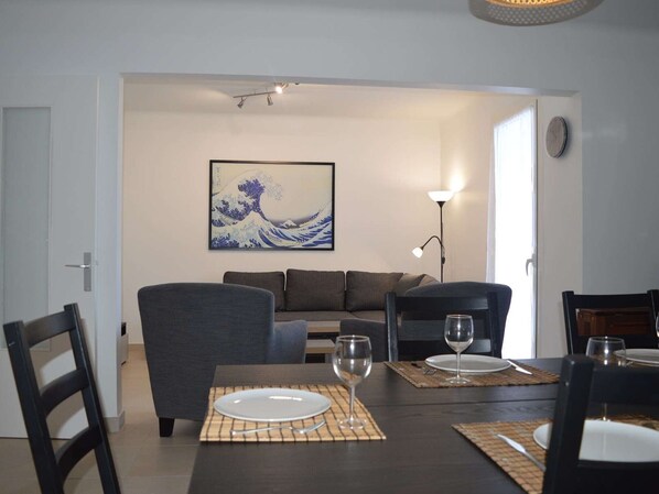 A dining area with a table set for two, a sofa, and a painting of a wave on the wall.
