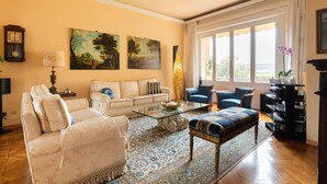 A living room with a beige sofa, a blue armchair, a glass coffee table, and a patterned rug. There are two paintings on the wall, a clock, and a lamp. - Luxury apartment next to the river (Firenze)