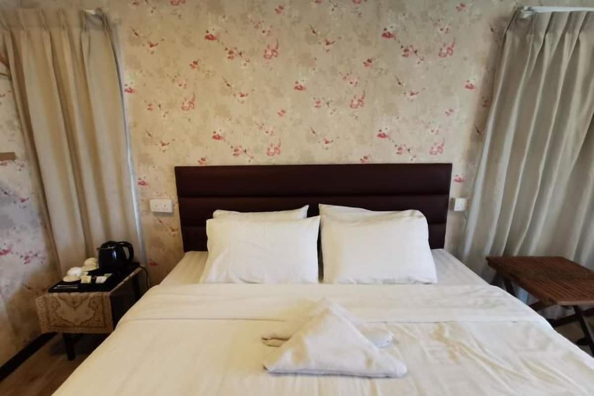 A hotel room with a bed, pillows, a nightstand with a kettle, and a small table.