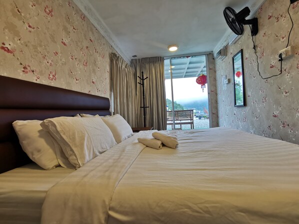 Une chambre d’hôtel avec un grand lit, une tête de lit, une fenêtre avec des rideaux, un ventilateur sur pied et une vue sur l’extérieur.