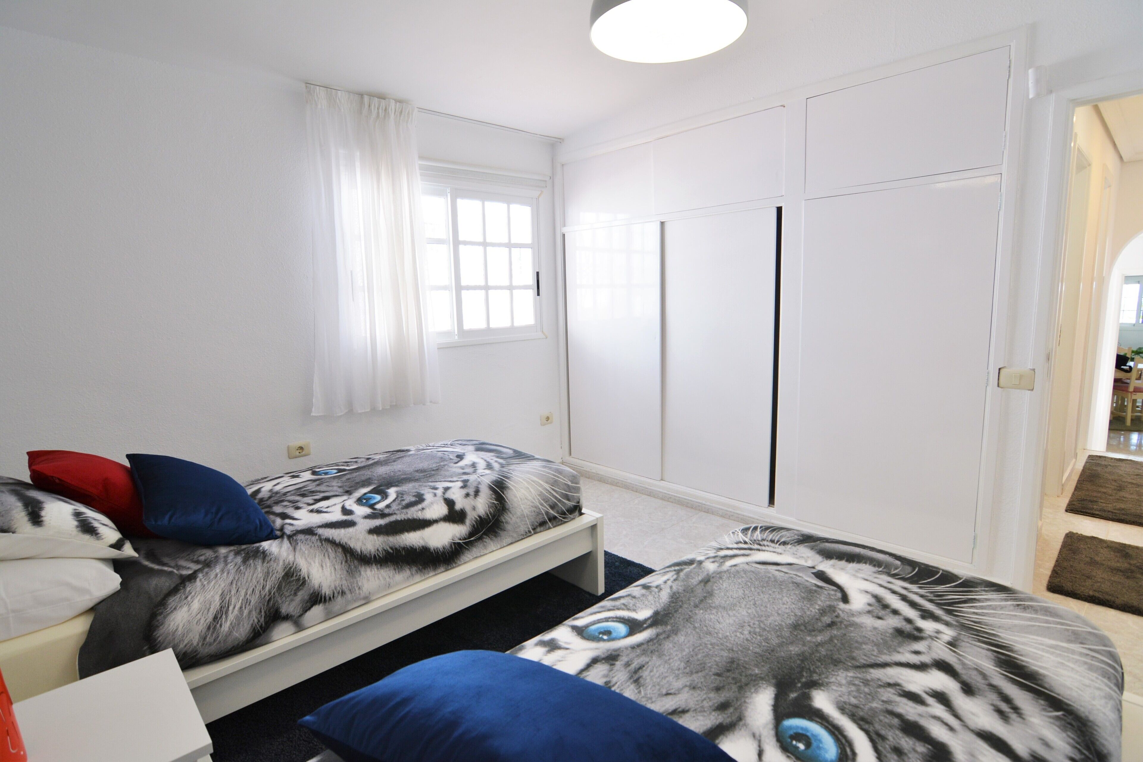A bedroom with two beds, white walls, a window with curtains, and a wardrobe.