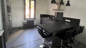 Eine moderne Küche mit einer zentralen Kochinsel, Barhocker, Pendelleuchten und Blick ins Freie durch ein Fenster.
