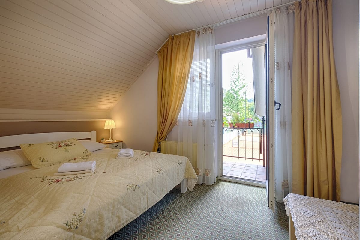 A bedroom with a bed, a bedside table with a lamp, a window with curtains, and a view of a garden.