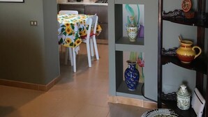 Un comedor con una mesa cubierta con un mantel de girasoles, rodeada de sillas. Hay estantes con varios jarrones de cerámica y un tazón decorativo en el suelo.