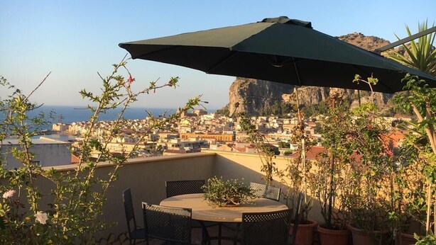Un patio con mesa y sillas, rodeado de plantas en macetas, con vista a una ciudad costera y un acantilado.
